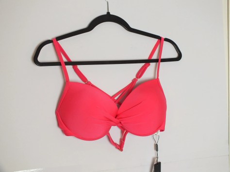a vibrant pink bikini top is hanging from a black hanger against a plain white wall. The bikini top features a twisted knot detail in the center of the bust, with thin straps that go over the shoulders and connect to a criss-cross back. The fabric appears to be a smooth, slightly shiny material, characteristic of swimwear. A small white tag is dangling from the side of the bikini. The lighting is even, casting minimal shadows, and the focus is sharp on the garment.