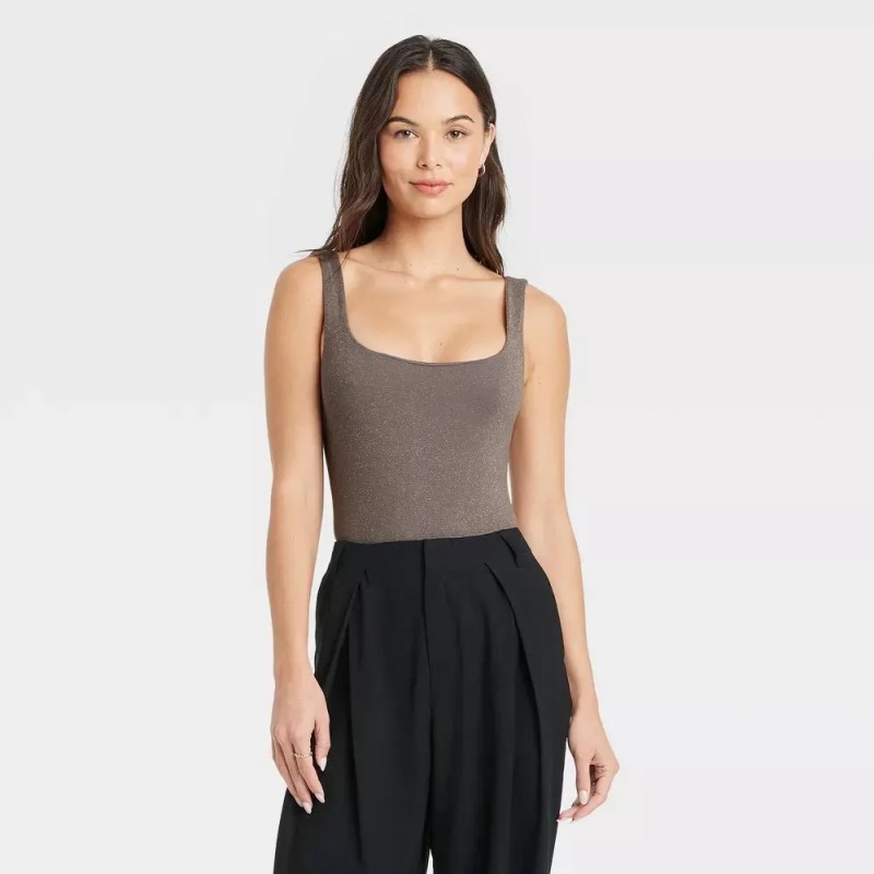 A young woman with long, dark, wavy hair stands facing the camera. She is wearing a taupe, sleeveless, square-neck tank top and high-waisted, pleated, black trousers. The background is a plain, light gray studio backdrop. The lighting is even and highlights the woman's features and the textures of her clothing.