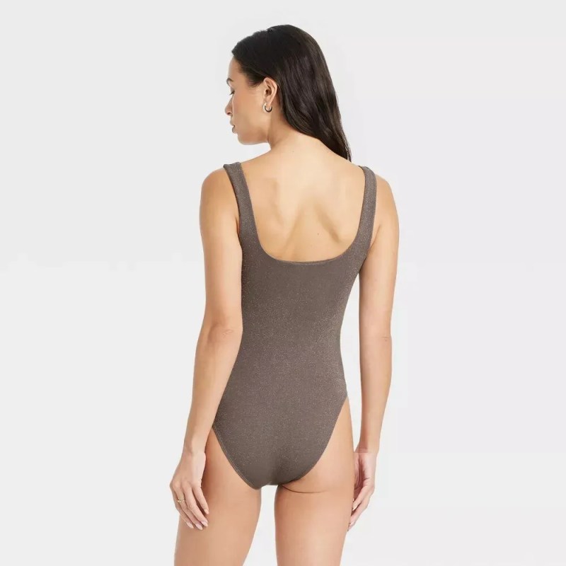 A woman with dark hair, seen from the back, stands against a plain white background. She is wearing a form-fitting, ribbed, taupe-colored one-piece swimsuit with a square neckline. The swimsuit has thick shoulder straps and a high-cut leg opening. Her skin tone is fair. The lighting is soft and even, with no harsh shadows. The image is a studio shot, focusing on the garment.