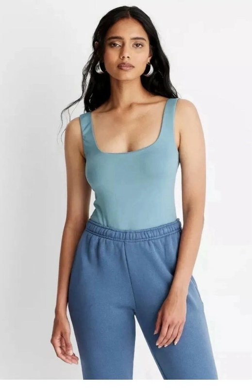 A medium shot portrait of a young Indian woman with long, dark, wavy hair and brown eyes. She is wearing a light teal ribbed square-neck tank top and loose, dark teal sweatpants. She has on large, silver hoop earrings. The woman is standing against a plain white background, with soft studio lighting. Her gaze is directed forward.